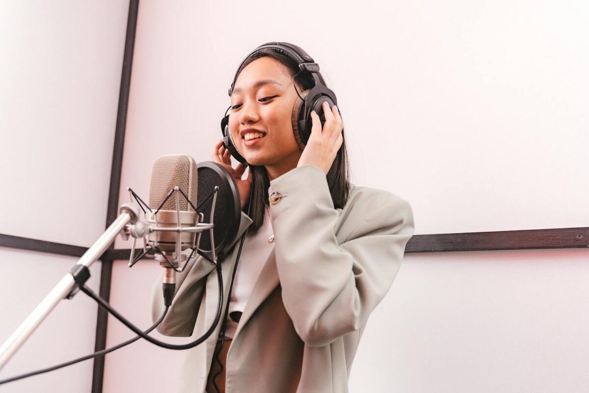Asian woman wearing headphones sings into a condenser microphone in a studio.